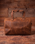 A brown distressed leather duffel bag with top handles, resting on a rustic wooden surface against a wooden plank background. The bag features the brand name '1847 Antiques' embossed on the front.