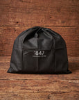 A black drawstring storage bag with the white printed text '1847 LEATHER EIRE' on the front, resting on a rustic wooden surface against a matching wooden background.