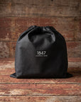 A black drawstring storage bag with white printed text reading '1847 LEATHER & EIRE' centered on the front, placed on a rustic wooden surface against a weathered wood plank background.