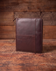 A dark brown leather zip-around document folder or padfolio cover standing upright on a rustic wooden table against a matching wood plank background.