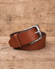 slim suit belt made of full grain vegetal leather in chestnut brown tan colour with polished brass style metal buckle on a rustic wooden table
