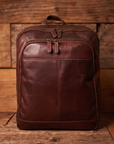 A dark brown leather backpack standing upright against a rustic wooden plank background.