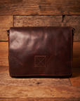 A dark brown leather messenger bag with a distressed finish, featuring a square stitched patch on the front, set against a rustic wooden plank background.