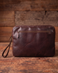 A dark brown leather laptop sleeve or document pouch with a wrist strap, resting on a rustic wooden surface against a matching wood plank background.