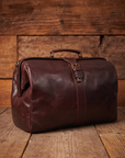 A dark reddish-brown doctors bag style vintage tan leather duffel bag with a top handle and buckle strap closure, set against a rustic wooden plank background.