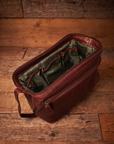A dark reddish-brown leather toiletry bag with an olive green interior, shown open on a rustic wooden plank floor.