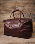 A dark reddish-brown leather duffel bag with double top handles and buckle details, displayed on a rustic wooden plank surface against a matching wood plank background.
