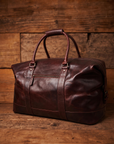 A dark reddish-brown leather duffel bag with double top handles and a front zipper pocket, photographed against a rustic wooden plank background.