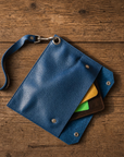 A blue textured leather pouch with a matching wrist strap and silver-tone hardware, shown open on a rustic wooden surface. Card slots reveal yellow, green, and brown cards.