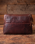 A dark brown distressed leather pouch bag with a front zippered pocket, resting on a rustic wooden surface with a matching wooden plank background.