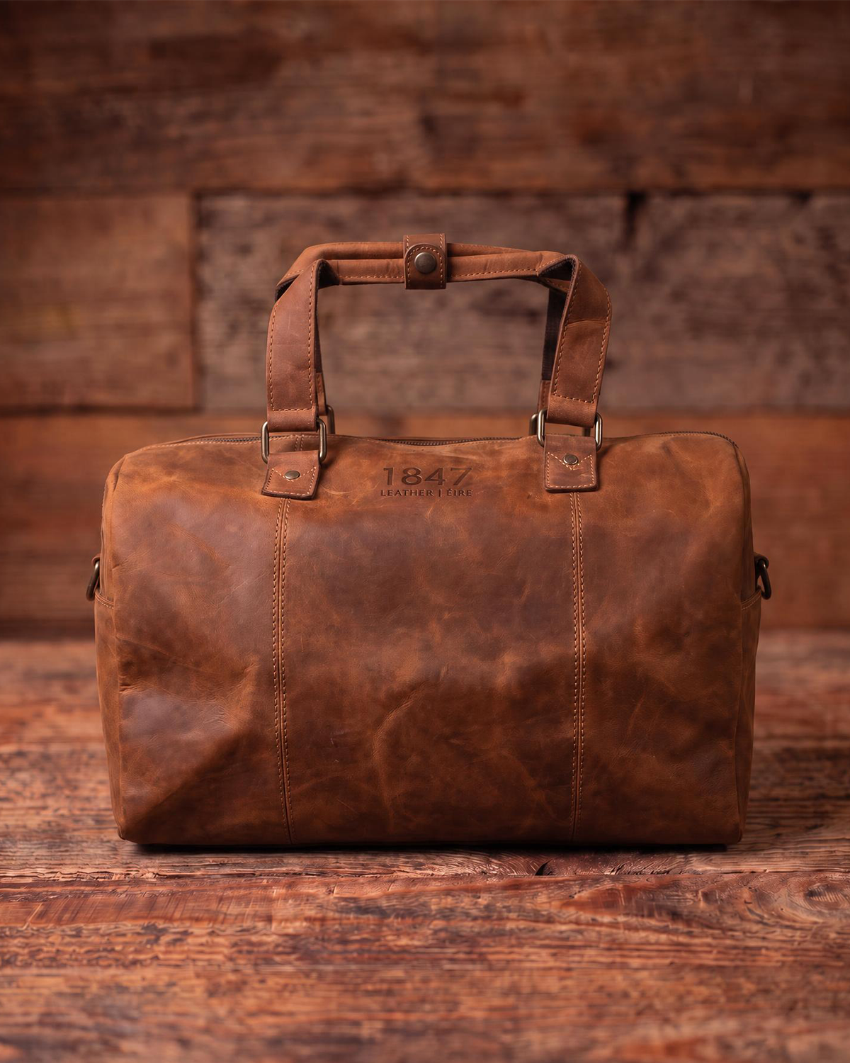A brown distressed leather duffel bag with top handles, resting on a rustic wooden surface against a wooden plank background. The bag features the brand name '1847 Antiques' embossed on the front.