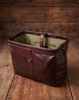 A dark reddish-brown leather doctors style weekender bag with a zippered interior and olive green lining, detachable strap. displayed on a rustic wooden plank floor against a matching wood plank background.