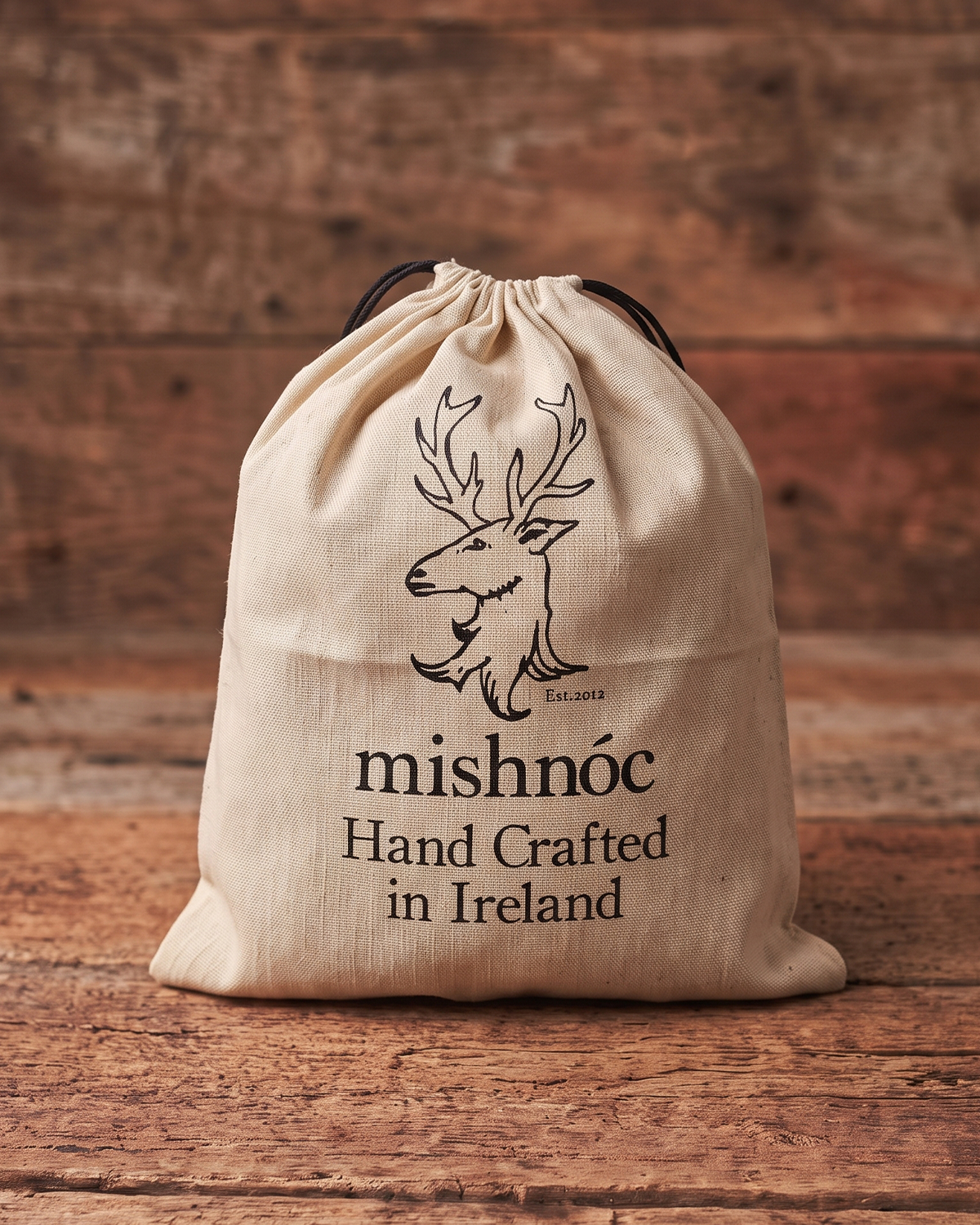 A beige recycled cotton drawstring dust bag for belt with black printed text reading 'mishnóc Hand Crafted in Ireland' and a deer logo, photographed on a rustic wooden surface with a wood plank background.