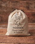 A beige recycled cotton drawstring dust bag for belt with black printed text reading 'mishnóc Hand Crafted in Ireland' and a deer logo, photographed on a rustic wooden surface with a wood plank background.