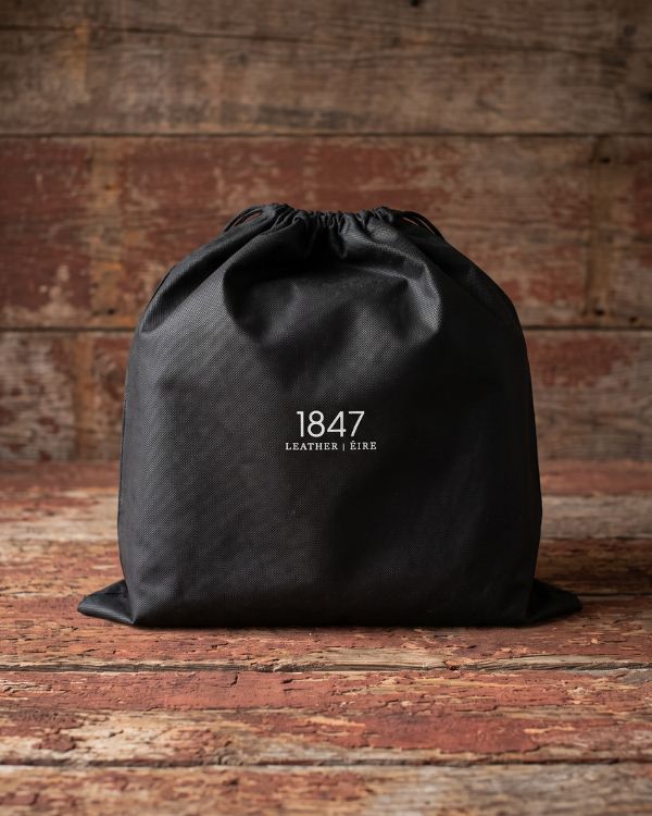A black drawstring storage bag with white printed text reading '1847 LEATHER &amp; EIRE' centered on the front, placed on a rustic wooden surface against a weathered wood plank background.