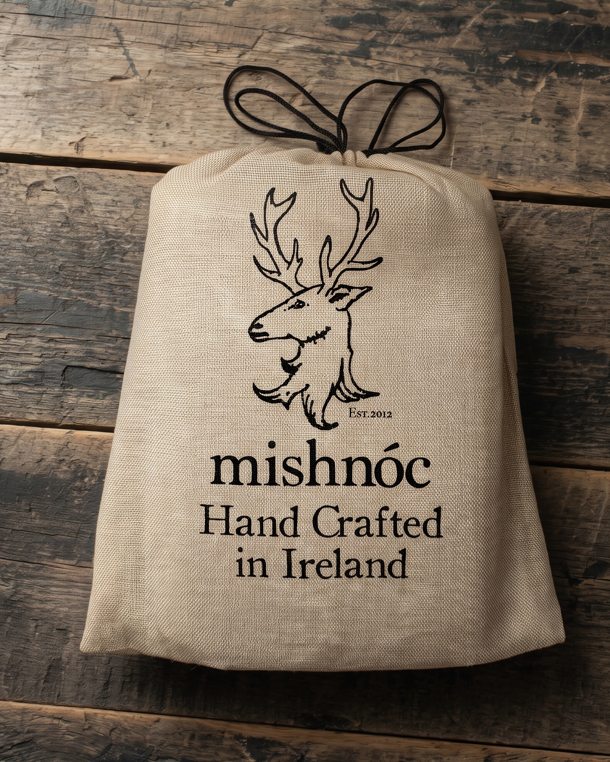 A beige canvas drawstring sack with a black printed deer head logo and the text 'mishnóc Hand Crafted in Ireland' laid flat on a rustic dark wood background.