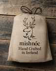 A beige canvas drawstring sack with a black printed deer head logo and the text 'mishnóc Hand Crafted in Ireland' laid flat on a rustic dark wood background.