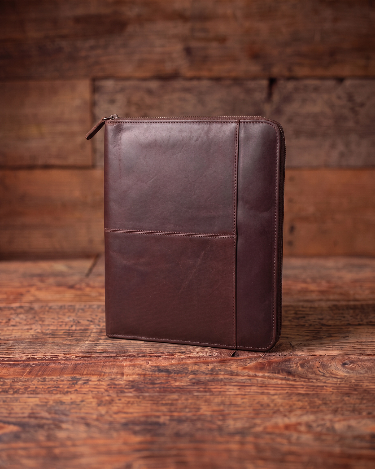 A dark brown leather zip-around document folder or padfolio cover standing upright on a rustic wooden table against a matching wood plank background.