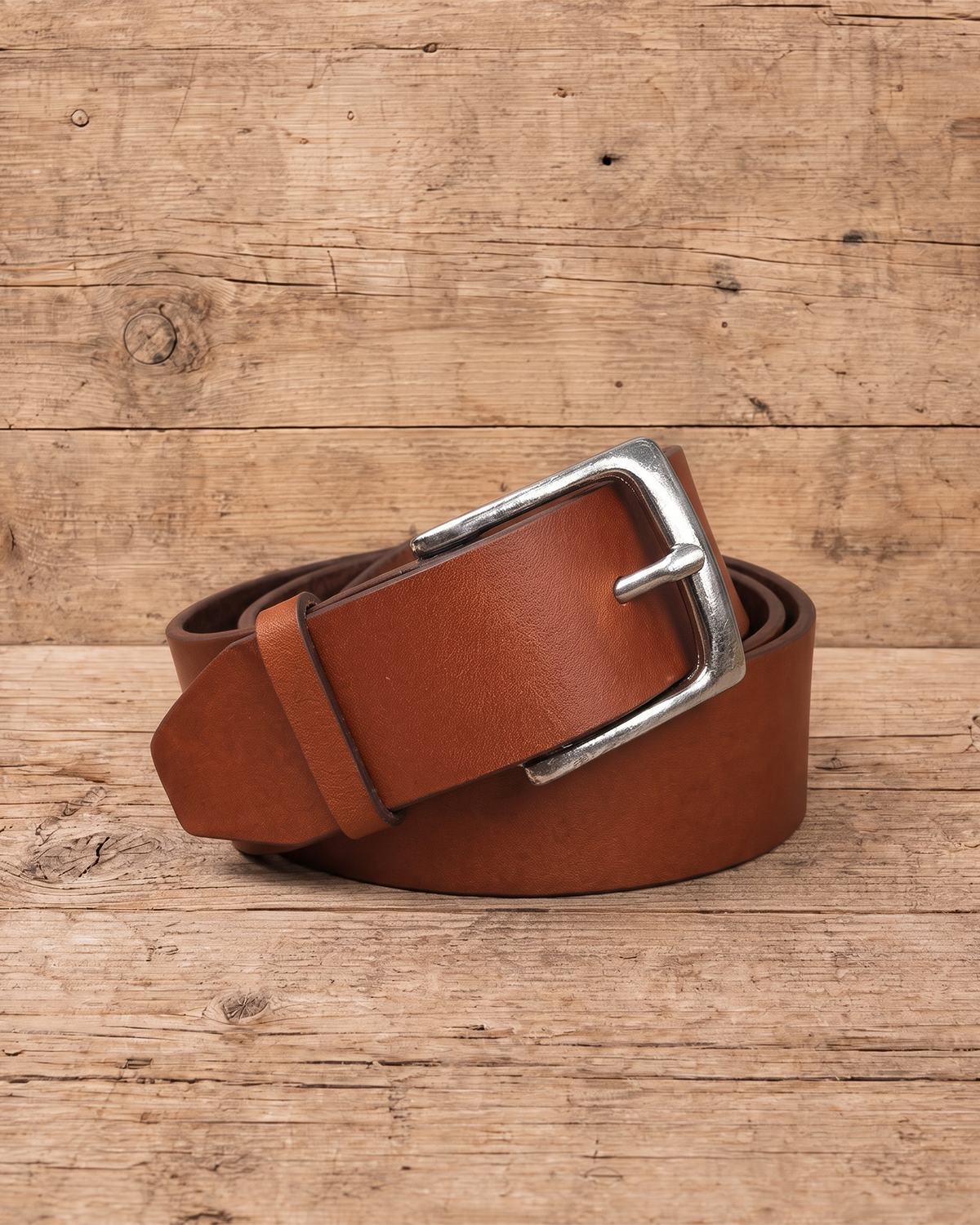 slim suit belt made of full grain vegetal leather in chestnut brown tan colour with polished brass style metal buckle on a rustic wooden table