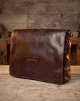 A dark brown distressed leather messenger bag with a front flap and a small stitched square patch, resting on a rustic wooden table with a blurred wood-paneled background.