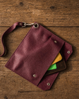A burgundy textured leather wristlet pouch with a metal clasp and strap, shown open on a rustic wooden background with cards partially visible inside.