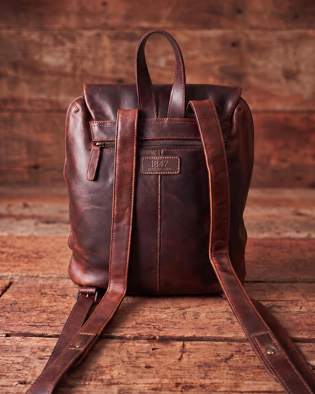 A dark brown distressed leather backpack shown from the back, featuring long shoulder straps, a top handle, and a zippered pocket, set against a rustic wooden plank background.
