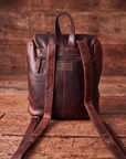 A dark brown distressed leather backpack shown from the back, featuring long shoulder straps, a top handle, and a zippered pocket, set against a rustic wooden plank background.
