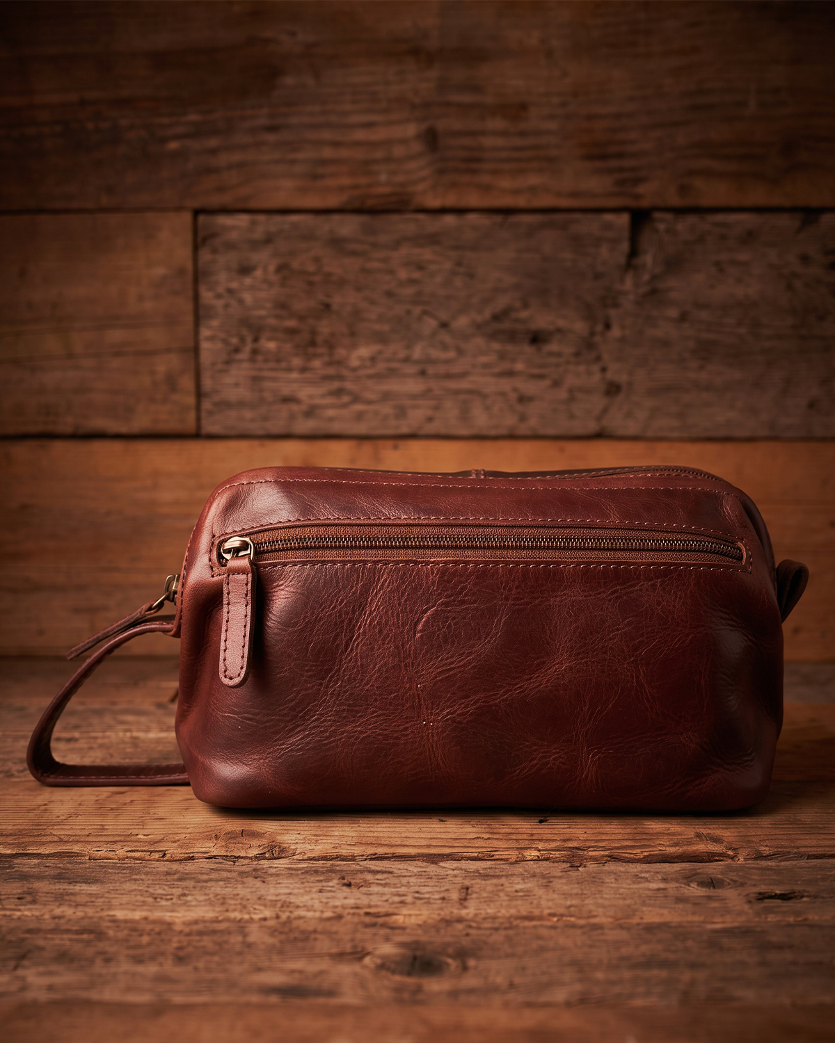 A brown leather crossbody pouch bag with a top zipper and wrist strap, displayed on a rustic wooden surface against a matching wood plank background.