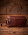 A brown leather crossbody pouch bag with a top zipper and wrist strap, displayed on a rustic wooden surface against a matching wood plank background.