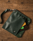 A dark green textured leather wristlet pouch with a metal clasp and strap, shown open on a rustic wooden surface with cards partially visible inside.