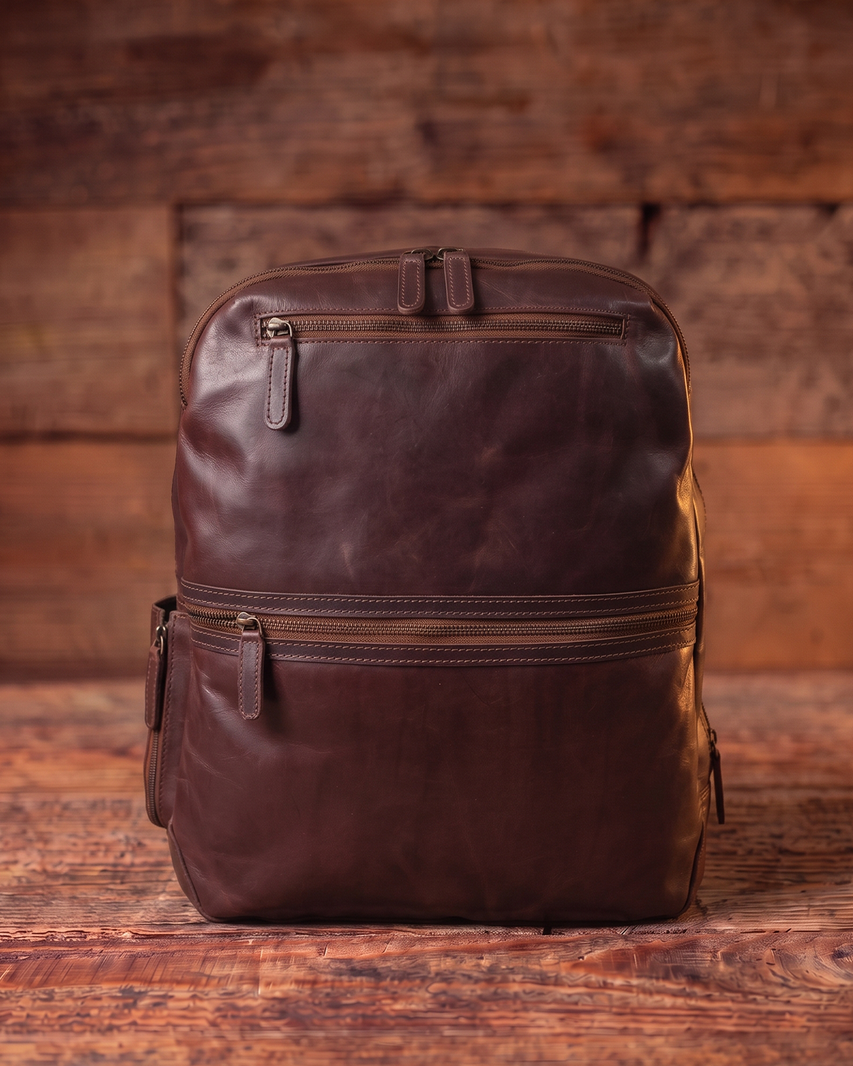 A dark brown leather backpack with multiple zippered compartments, shown standing upright against a rustic wooden plank background.