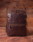 A dark brown leather backpack with multiple zippered compartments, shown standing upright against a rustic wooden plank background.