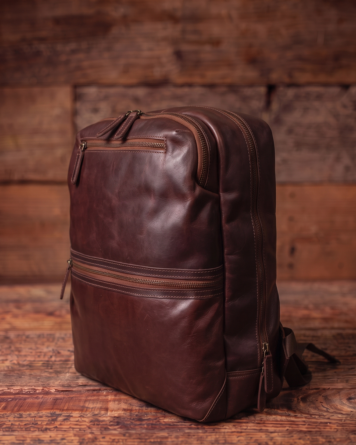 A dark brown distressed leather backpack with multiple zippered compartments, shown from a three-quarter angle on a rustic wooden surface with a matching wooden plank background.