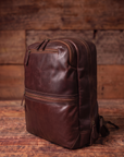 A dark brown distressed leather backpack with multiple zippered compartments, shown from a three-quarter angle on a rustic wooden surface with a matching wooden plank background.