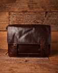 A dark brown leather satchel or laptop messenger bag with brass-tone buckles, displayed on a rustic wooden plank surface against a matching wood plank background.