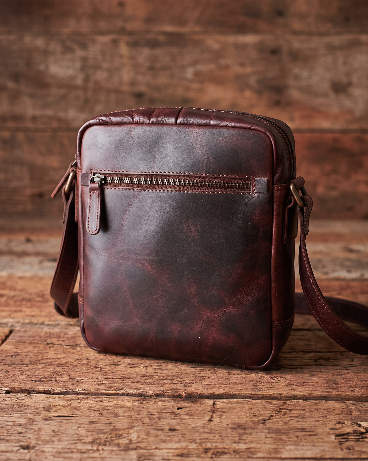 A dark reddish-brown leather crossbody bag with a front zippered pocket, resting on a rustic wooden surface against a matching wood plank background.