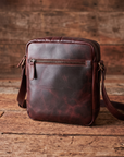 A dark reddish-brown leather crossbody bag with a front zippered pocket, resting on a rustic wooden surface against a matching wood plank background.