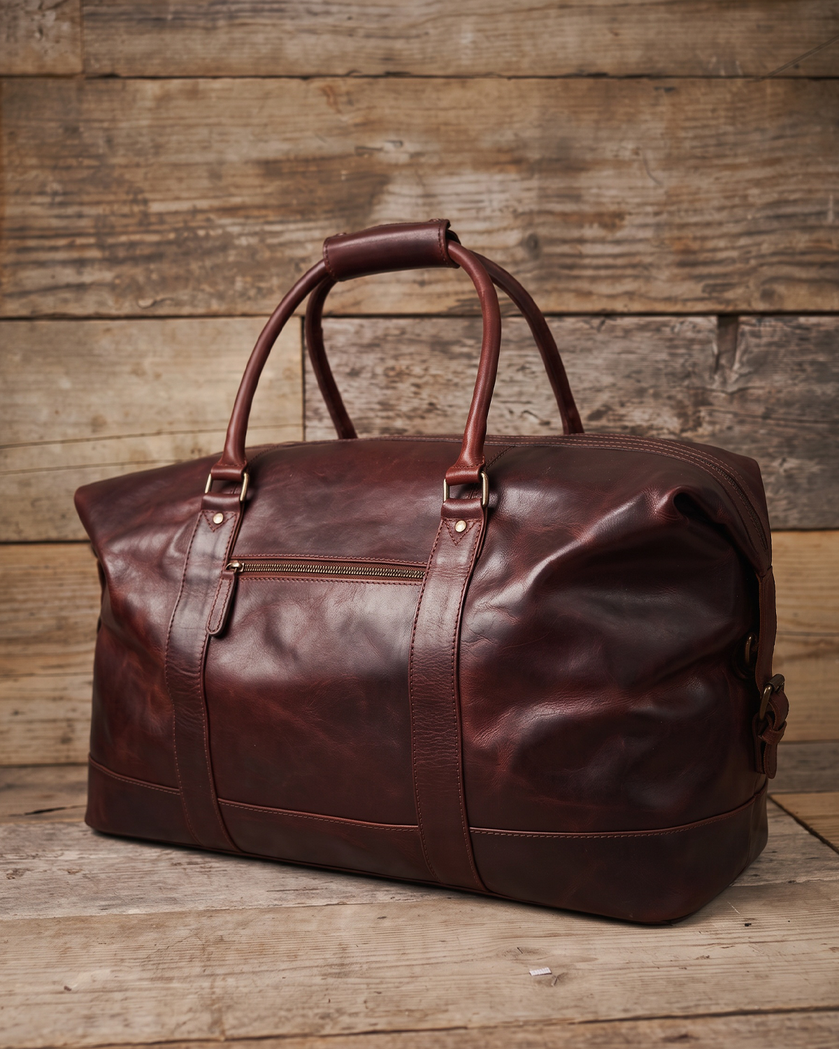 A dark reddish-brown leather duffel bag with double top handles and buckle details, displayed on a rustic wooden plank surface against a matching wood plank background.