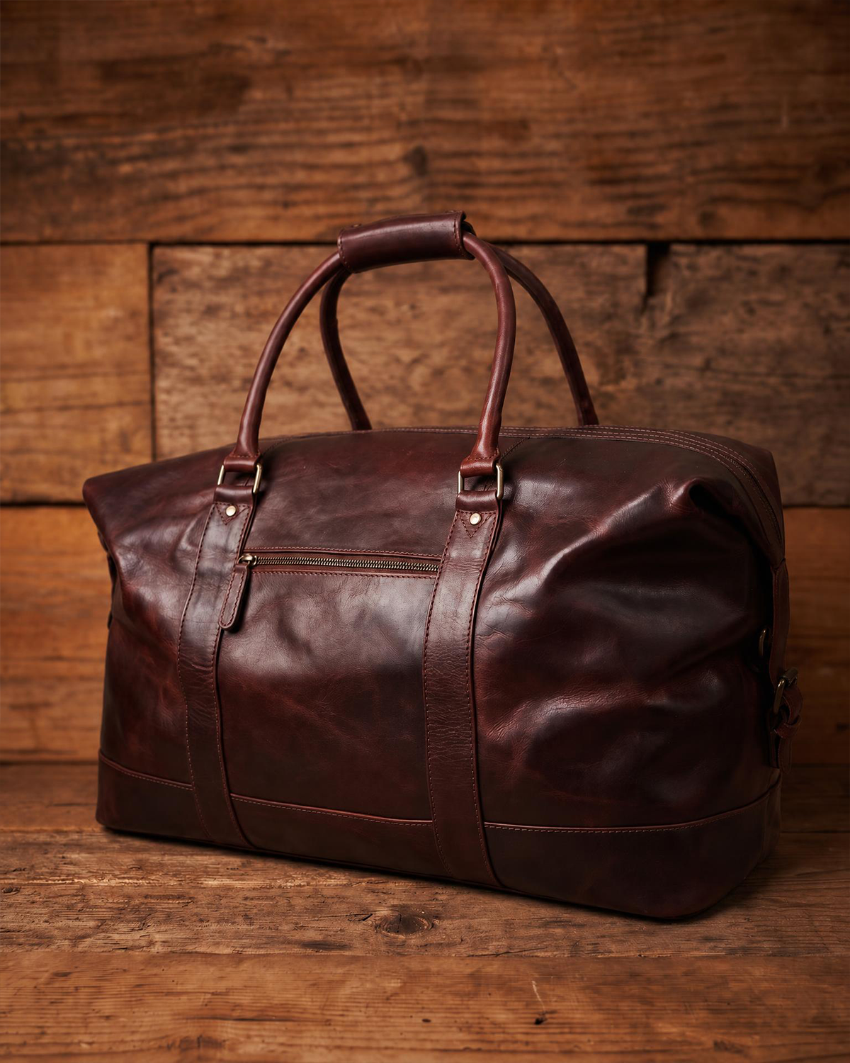 A dark reddish-brown leather duffel bag with double top handles and a front zipper pocket, photographed against a rustic wooden plank background.