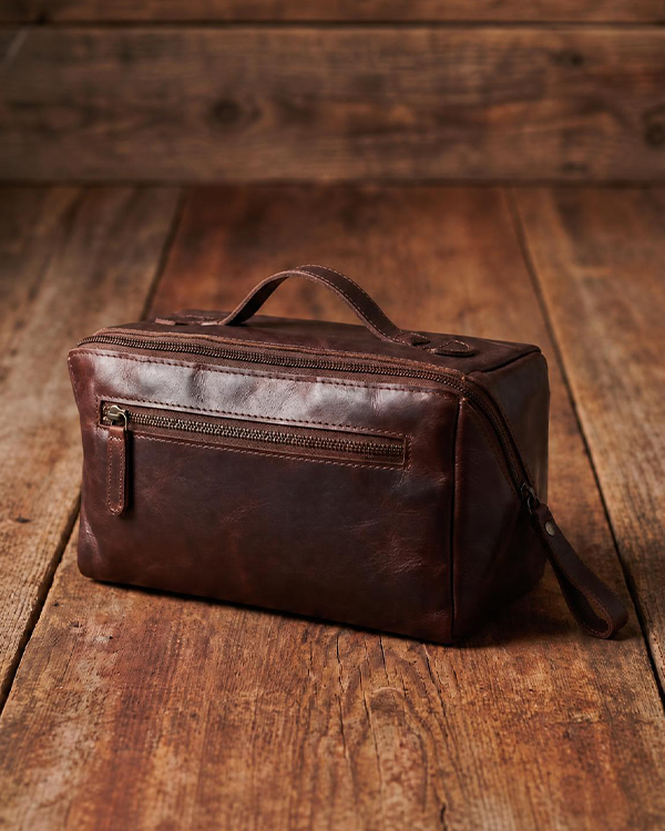 A dark brown leather toiletry bag with a top handle and front zipper pocket, resting on a rustic wooden plank surface.