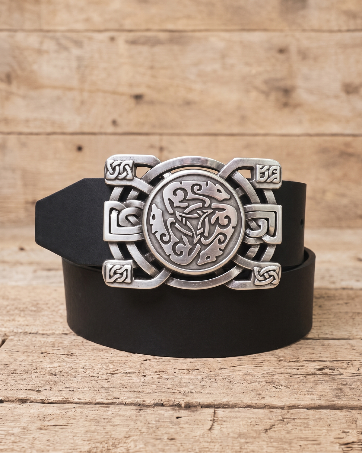 A black leather belt with an ornate silver-toned Celtic knot buckle, coiled on a rustic wooden surface.