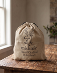 A beige canvas drawstring bag with a black printed deer logo and the text 'mishnóc Hand Crafted in Ireland' sits on a rustic wooden table in a softly lit indoor setting.