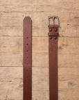 slim suit belt made of full grain vegetal leather in chestnut brown tan colour with polished brass style metal buckle on a rustic wooden table