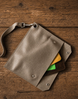 A textured leather pouch in a taupe-beige color, shown from a top-down view on a rustic wooden background. The pouch has a matching leather strap with a silver metal clasp, snap buttons, and is shown holding yellow and green card-like inserts.