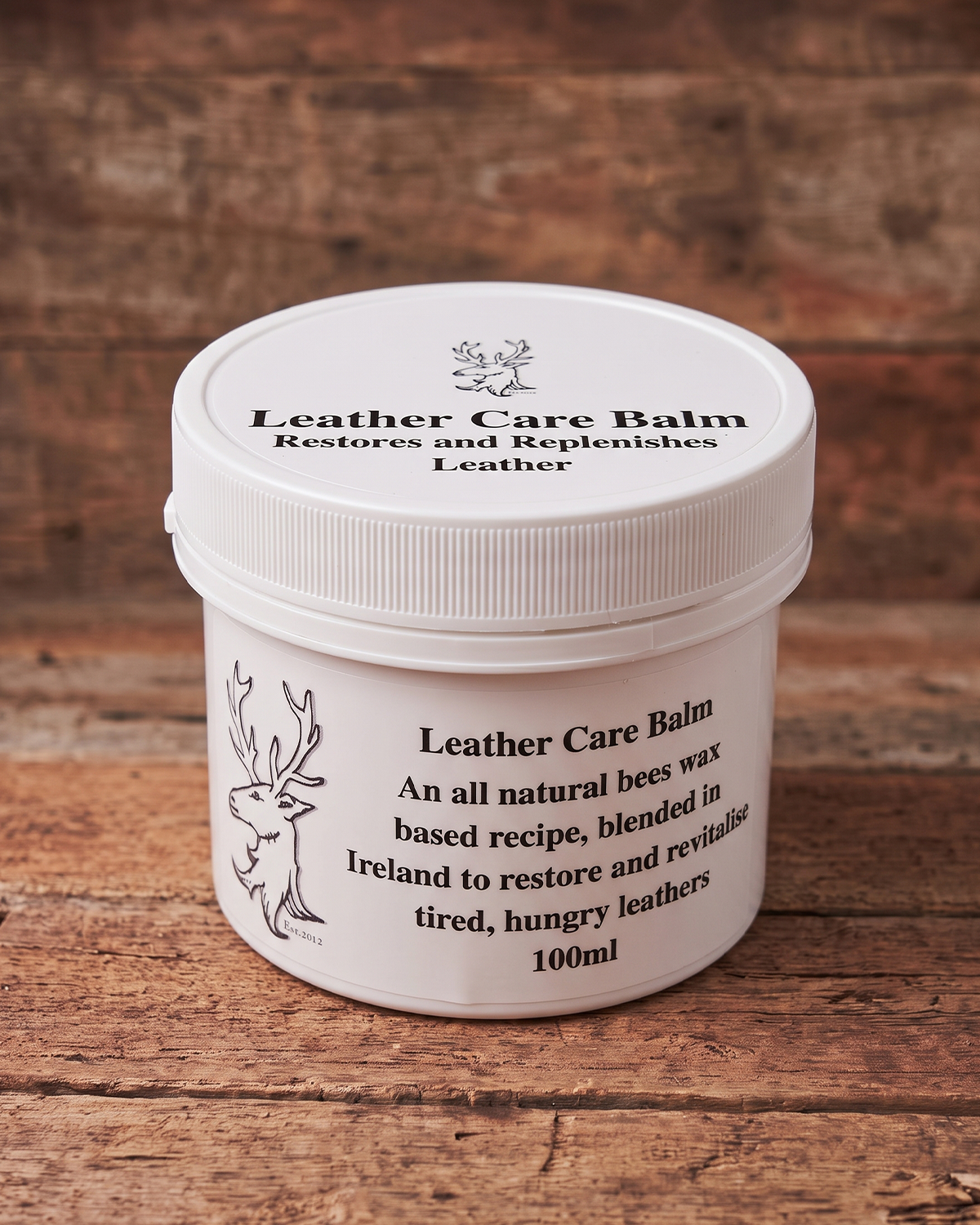A white plastic jar of Leather Care Balm with a ribbed screw-top lid, placed on a rustic wooden surface. The label features black text and a simple black line drawing of a deer.